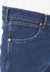 Ciemnoniebieskie dżinsy z denimu o dopasowanym kroju, z pomarańczowym przeszyciem, przednimi kieszeniami i detalem w postaci miedzianego guzika na pasie.