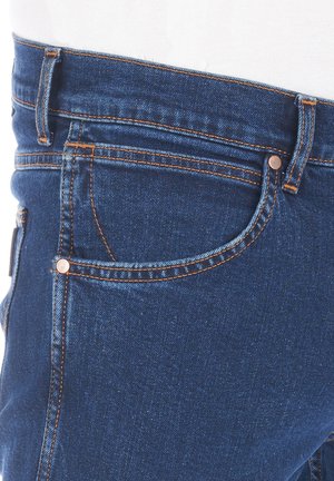 Jeans in denim blu con una vestibilità stretta, cuciture in rame e una piccola tasca frontale. Il tessuto ha una texture liscia con una leggera lucentezza.