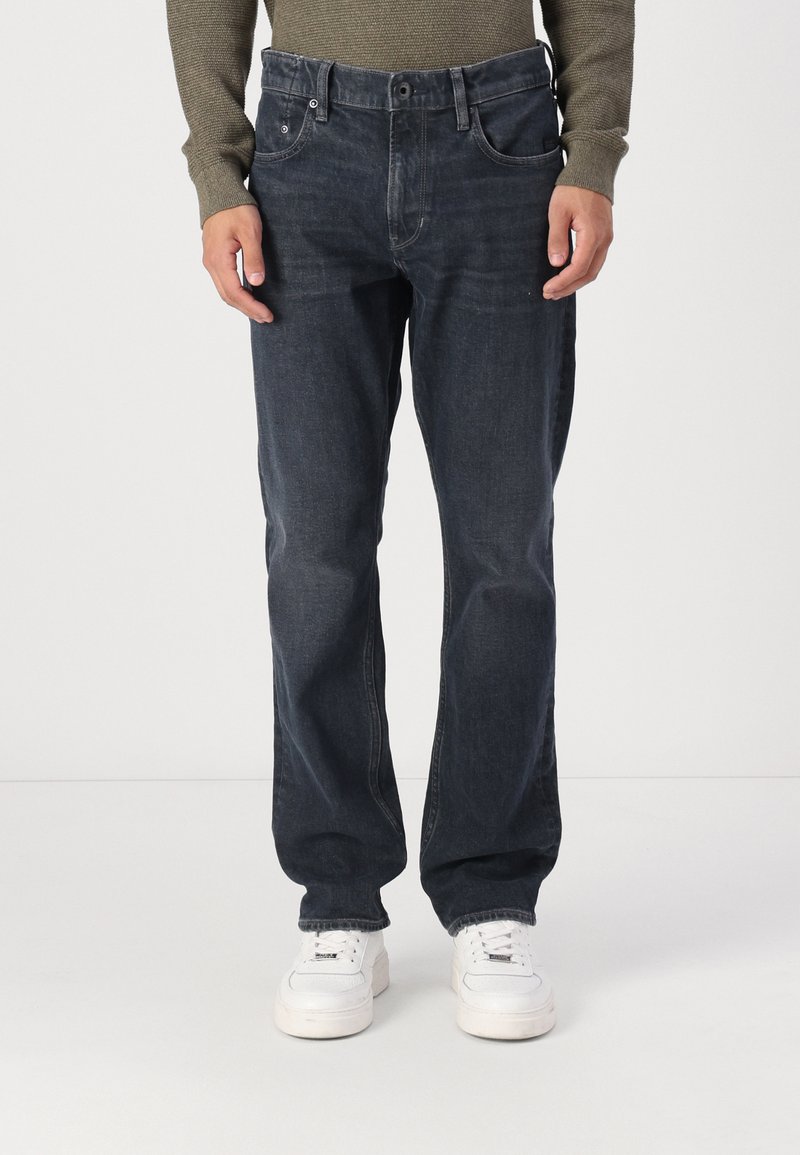 Mørkeblå denimjeans med rett passform, fem lommer og lett falming. Kombinert med hvite sneakers på en enkel bakgrunn.