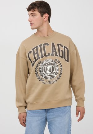 Felpa beige con una grande stampa "CHICAGO" e un emblema con dettagli verde oliva, polsini a costine e una vestibilità comoda. Indossata con jeans azzurri chiari.