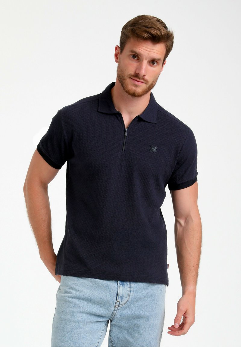 Marineblå polo skjorte med kort lynlås, struktureret stof, krave og korte ærmer. Bæret med lyseblå jeans, der har et lille logo på brystet.