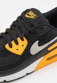 Nike-sneaker med överdel i svart mocka och mesh, gula gummidetaljer, grå swoosh, strukturerade snören och vit mellansula med orange detaljer.