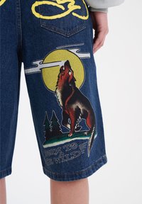 Denim shorts, joissa on susigrafiikka, joka ulvoo keltaista kuuta kohti, puiden ympäröimänä, ja teksti "BORN TO BE WILD" valkoisena.