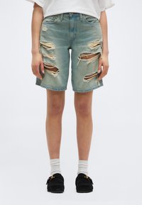 Άτομο που φοράει ξεθωριασμένα σορτς από τζιν σε ανοιχτό μπλε με μεγάλα σκισίματα, λευκές κάλτσες, μαύρα παντόφλες χωρίς κορδόνια και λευκό τοπ, σε απλό φόντο.