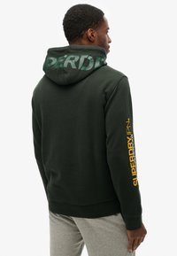 Felpa con cappuccio verde scuro, con un grande logo sul cappuccio, testo giallo sul manico e polsini a coste. Realizzata in morbido materiale misto cotone.