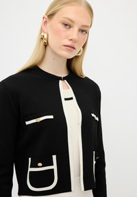 Cardigan nero con bordi bianchi, dotato di tasche frontali e dettagli con bottoni dorati. Texture liscia, design con colletto e dettaglio a forma di chiave sul collo.