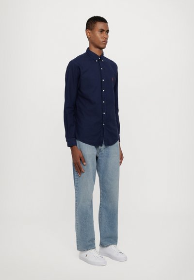 Marineblauwe overhemd met kraag, gecombineerd met lichtblauwe rechttoe rechtaan spijkerbroek en witte sneakers. Het shirt heeft een klein logo op de borst.