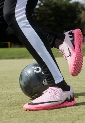 Jugador con pantalones blanco y negro y zapatillas rosas controla un balón de fútbol negro en un campo de césped.