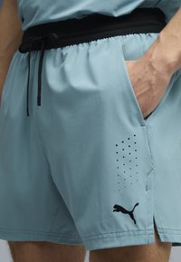 Άνδρας που φοράει ανοιχτό μπλε αθλητικό σορτς με μαύρο κορδόνι, με το χέρι στην τσέπη και μαύρο λογότυπο Puma στο αριστερό πόδι.