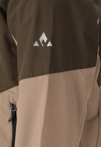 Vandafvisende jakke i mørkebrun og beige. Sølv logo på brystet. Har en lynlåslomme på venstre side, glat tekstur.