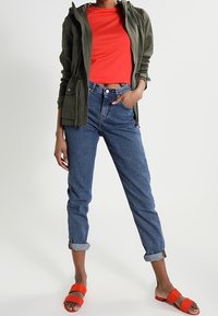 Olivgrön huvjacka, röd åtsittande t-shirt, högmidjade blå denimjeans, uppvikta vid anklarna, tillsammans med oranga slip-on sandaler.