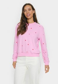 Roze sweatshirt met rode hartpatronen, ronde hals en lange mouwen. Zacht materiaal met geribde manchetten en zoom, gecombineerd met witte broek.