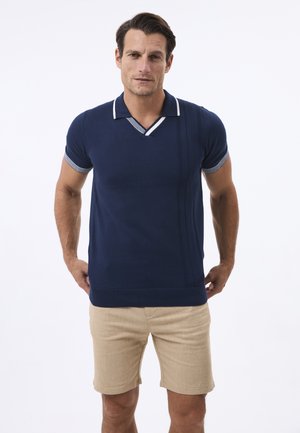 Herren Poloshirt in Marineblau mit V-Ausschnitt und weißem Streifenakzent, mit kurzen Ärmeln und vertikalen Rippdetails auf einer Seite. Kombiniert mit beigen Shorts.