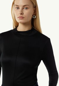 Czarna bluzka z długim rękawem, o wysokim dekolcie i dopasowanym kroju. Gładka tekstura z centralnym szwem. Modelka nosi srebrne kolczyki w kształcie kół.