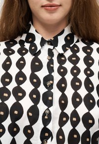 Nahaufnahme einer Person, die ein schwarz-weiß gemustertes, mit Knöpfen versehenes Hemd mit einem blattähnlichen Design und einem spitzen Kragen trägt.