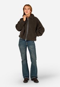 Pruun fliistee äkki-jakk, millel on krae, elastsed mansetid ja kaks eesmist taskut, kombineeritud flöödidest siniste teksade ja tumedate kingadega.