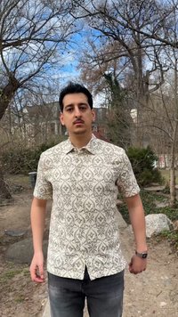 Homme debout en plein air sur un chemin de terre, portant une chemise à manches courtes à motifs et un jean foncé, avec des arbres dénudés et des bâtiments en arrière-plan.