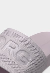 Gros plan sur la sangle d'une sandale slide lavande avec un tissu texturé et les lettres embossées "RG" sur un fond blanc uni.