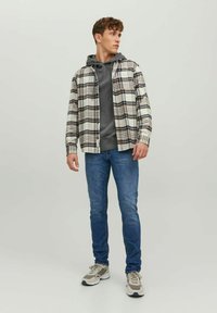 Camisa de franela a cuadros en blanco, beige y marrón sobre una sudadera con capucha gris. Jeans de mezclilla en azul claro. Zapatillas deportivas en gris y blanco.