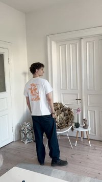 Λευκό βαμβακερό μπλουζάκι με πορτοκαλί γραφικό σχέδιο στην πλάτη με κείμενο "ΧΩΡΙΣ ΡΕΠΟ." Συνδυάζεται με φαρδιά σκούρα μπλε τζιν και μαύρα παπούτσια.