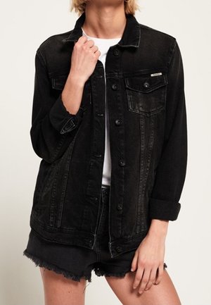 Osoba ubrana w czarną kurtkę z dżinsu na białą koszulę oraz czarne postrzępione szorty z dżinsu, trzymająca jedną ręką kołnierz kurtki.