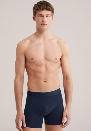Jeune homme en bonne forme portant un boxer bleu marine avec une ceinture "FUNDAMENTALS", debout devant un fond clair uni, face à l'avant.