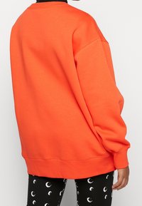 Ljusorange sweatshirt med avslappnad passform, ribbat halsband och långa ärmar, kombinerad med svarta leggings med ett vitt halvmånemönster.