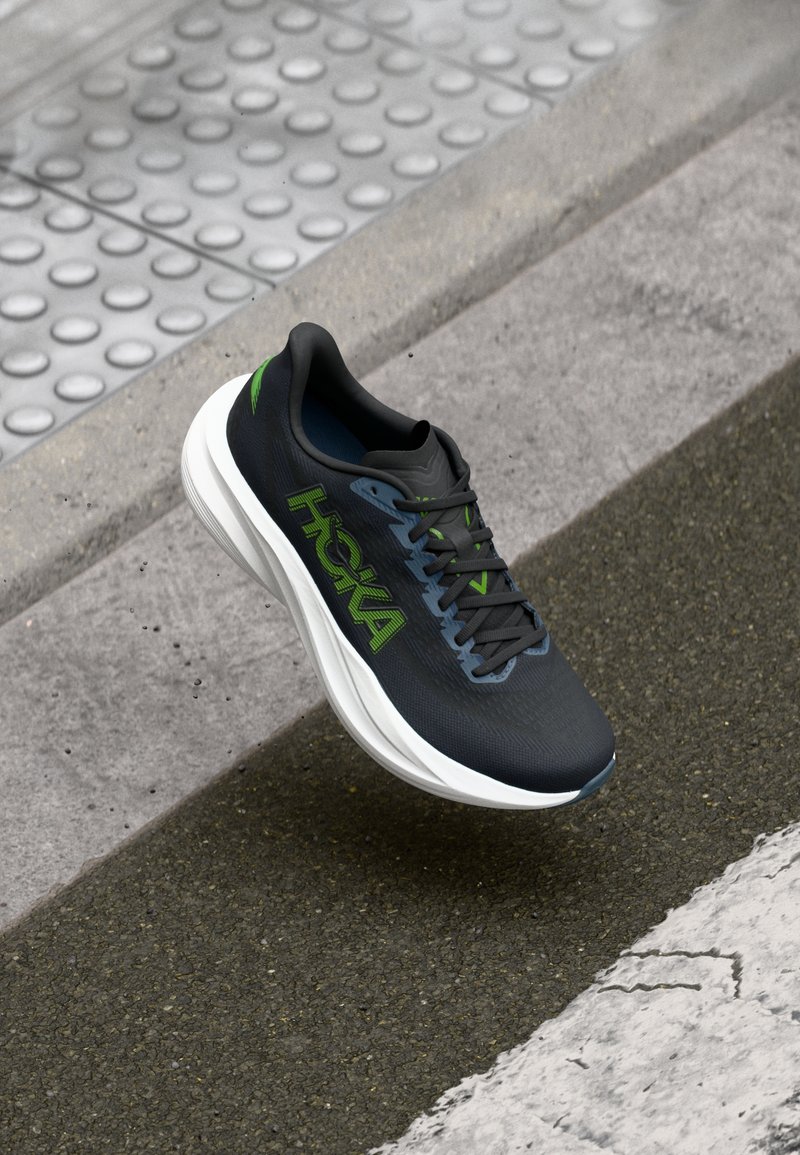 Black and green Hoka running shoe mid-air above textured pavement and sidewalk curb with tactile paving in background.