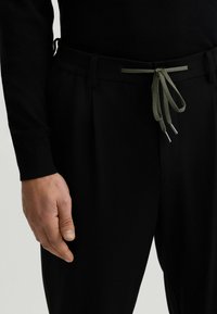 Schwarze Hose mit elastischem Bund, flachem Vorderteil und verstellbarem olivgrünem Kordelzug mit Metallschnüren.