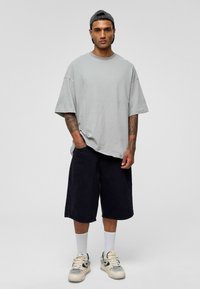Lichtgrijze oversized t-shirt, zwarte denim shorts, witte sokken en tan sneakers. Heeft een relaxte pasvorm met korte mouwen en zichtbare tatoeages.