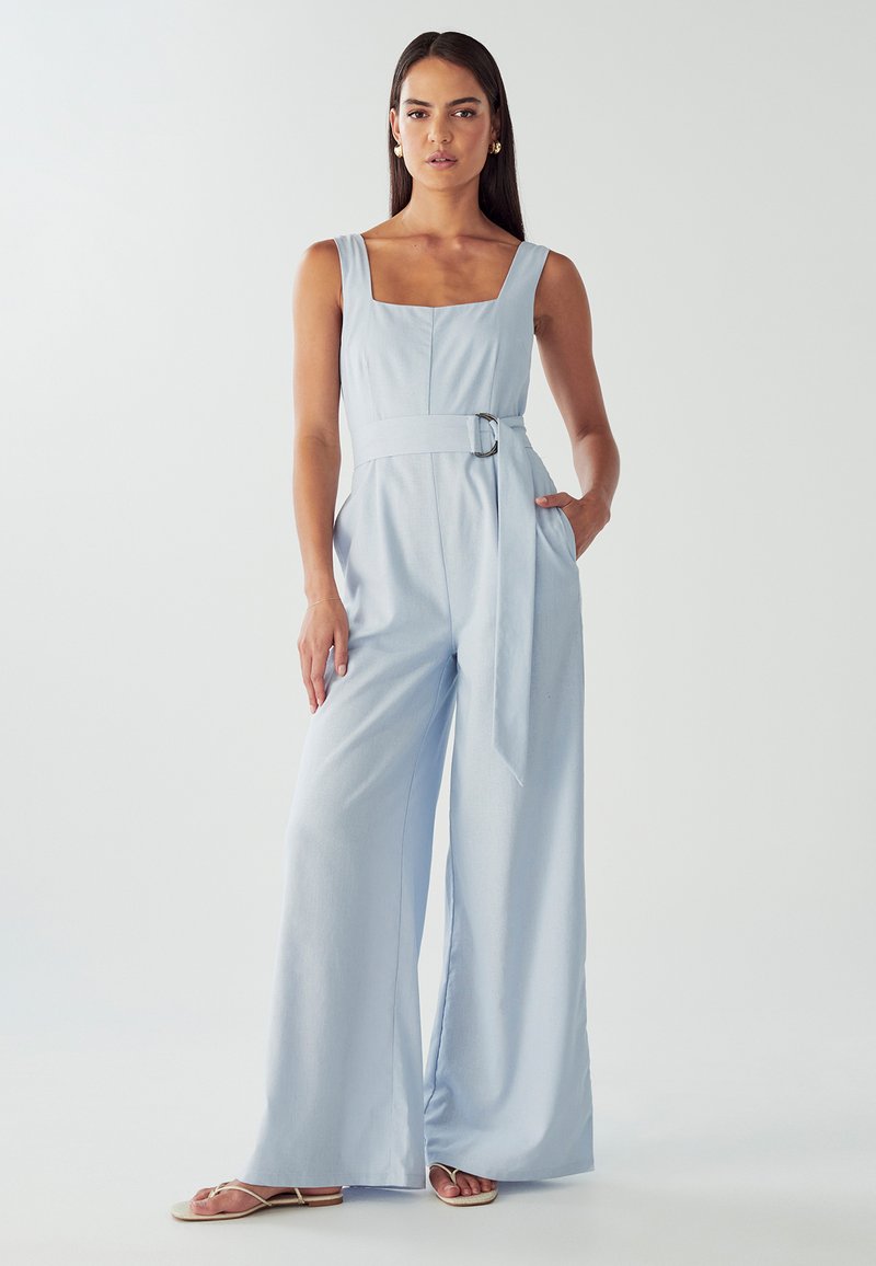Combinaison légère bleu clair avec jambes larges, décolleté carré et taille cintrée ornée d'une boucle en argent. Le tissu a une texture lisse.