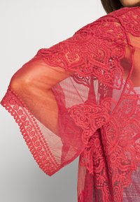 Läpinäkyvä punainen pitsikimono, jossa on monimutkaiset kukkakuviot, lovitetut reunat ja teksturoidut yksityiskohdat hihoissa ja reunoissa.