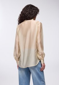 Blusa a maniche lunghe in beige chiaro, realizzata in tessuto trasparente, presenta un orlo arrotondato e polsini con bottoni. Indossata con jeans in denim blu chiaro.