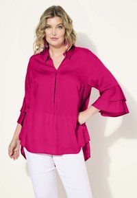 Fuchsia-Bluse mit V-Ausschnitt, drei Viertel langen Rüschenschößen und lässiger Passform. Glatter Stoff mit seitlichen Schlitzen, kombiniert mit weißen Hosen.