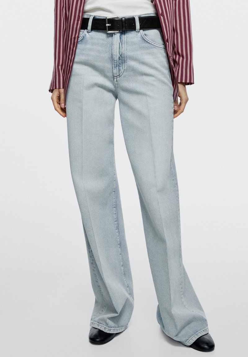 Femme portant un jean large bleu clair avec pli sur le devant, une ceinture noire, des chaussures noires, un haut blanc et une chemise rayée bordeaux.
