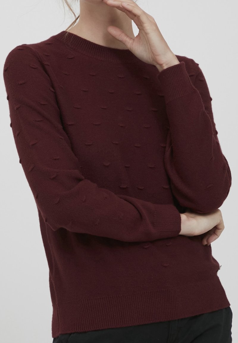 Personne portant un pull en maille texturée rouge foncé avec de petits points en relief, bras croisés, une main touchant le menton.