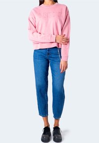Femme portant un sweat-shirt Levi's rose, un jean taille haute bleu avec des détails en chaîne, et des chaussures noires à lacets, debout devant un fond uni.