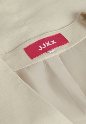 Beżowa tkanina o gładkiej teksturze; posiada czerwoną metkę z napisem "JUXX" w białym kolorze wszytą w dekolcie. Czysty design o prostym kroju.