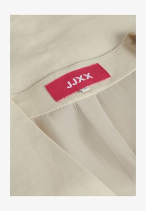 Bežinė medžiaga su lygiu paviršiumi; turi raudoną etiketę su "JUXX" balta tekstu, siuvama į apykaklę. Tvarkingas dizainas su paprastu kirpu.