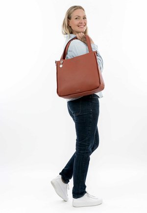 Braune, strukturierte Tote-Bag mit Reißverschluss, silberner Hardware und kurzen Griffen; von einer Person in lässiger Kleidung über der Schulter getragen.