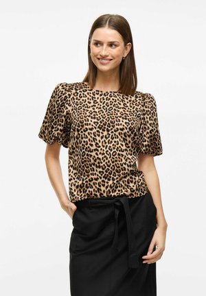 Leopardi-kuosiset paita silkkisessä kankaassa, jossa on puffihihat ja pyöreä pääntie, yhdistettynä mustiin hameisiin, kädet taskuissa.