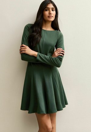 Robe verte à manches longues avec un corsage ajusté et une jupe évasée, fabriquée dans un tissu lisse, avec des détails de coutures et un col rond.