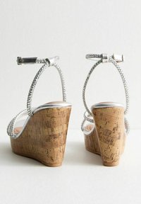 Sandalias de cuña plateadas con un tacón de corcho, tiras de rhinestone y una parte delantera transparente. La tira trasera es ajustable con un cierre metálico.