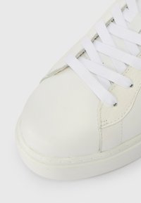 Balti ādas kedas ar noapaļotu purngalu, gludu tekstūru un baltām šņorēm. Iekļauj diskrētu izšuvumu akcentus un gumijas zoli.