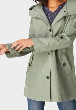 Femme ajustant la manche d'un imperméable à capuche vert clair avec poches boutonnées, portant un chemisier bleu et un jean.