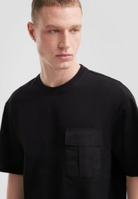 Czarna koszulka z bawełny, z teksturowaną kieszenią z klapką z przodu. Krótkie rękawy i okrągły dekolt. Gładki, casualowy design.