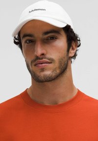 Gorra blanca con el logo de "lululemon", combinada con una camiseta de cuello redondo de color naranja. Presenta una tela suave y un escote redondo. Barba con textura natural.