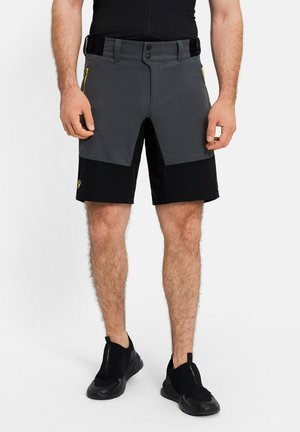 Mann trägt schwarz-grau geteilte Shorts mit seitlichen Reißverschlusstaschen und schwarzen Slip-On-Sportschuhen, steht vor weißem Hintergrund.