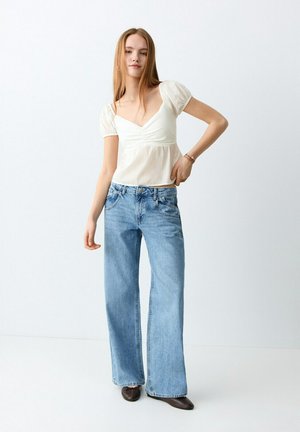 Mlada žena s dugom ravnom kosom, odjevena u bijelu bluzu s pufnastim rukavima, svijetloplave široke traperice i tamne cipele, stoji na jednobojnoj pozadini.