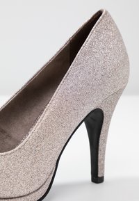 Zapato de tacón alto color rosa dorado brillante con puntera en almendra, forro interior suave y tacón negro afilado, que presenta una textura brillante.